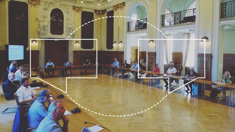 Es ist ein Foto eines größeren historischen Saals zu sehen. Mehrere Personen sitzen in diesem Saal an Tischen, alle Personen können sich sehen und sind einander zugewandt. Auf dem Foto wurde eine Grafik eingezeichnet, die zwei Sprechblasen mit einer gestrichelten Linie verbindet.