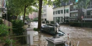 überflutete Straße und Auto, braunes ca. 50 cm tiefes Wasser, aus dem Mauern, Zäune, Bäume, mehrstöckige Häuser ragen, 