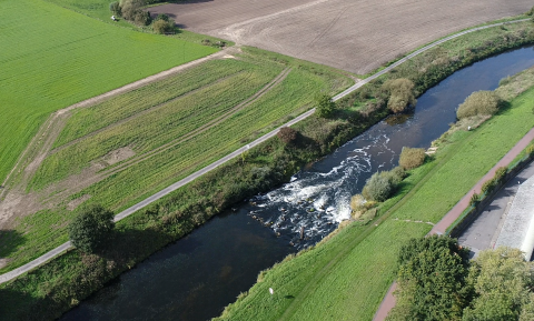 Das Luftbild zeigt das Gewässer mit dem Wehr (erkennbar am weiß aufschäumenden Wasser) in der Agrarlandschaft. Auf beiden Uferseiten wird das Gewässer von einem Weg bzw. einer Straße begleitet.
