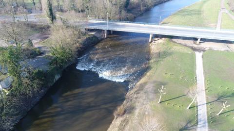 Das Luftbild zeigt das Wehr (erkennbar am weiß aufschäumenden Wasser). Über das Gewässer führt eine Straßenbrücke. Am rechten Ufer führt ein Weg flussbegleitend durch eine Wiesenlandschaft, rechts ist Baumbestand.