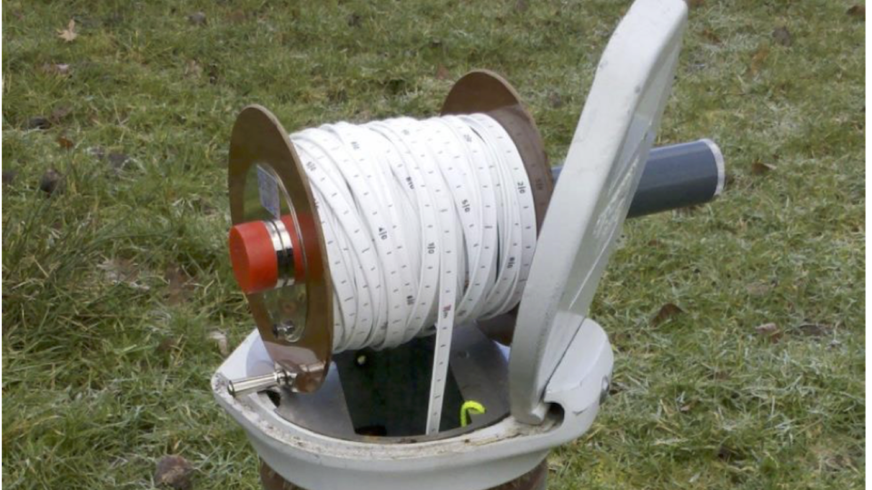 Auf dem Foto ist ein aufgerolltes Maßband zu sehen, dass in ein Rohr an einer Grundwassermessstelle hineinreicht.