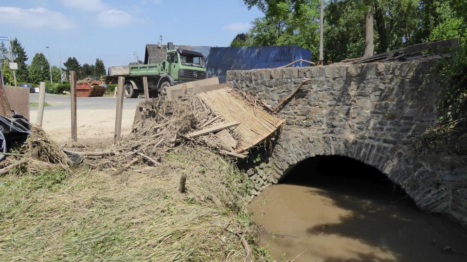 Steinbogenbrücke, Fluss, der schlammig-braunes Wasser führt, angeschwemmtes Holz und zerstörter Zaun am Ufer, Lastwagen, Müllcontainer